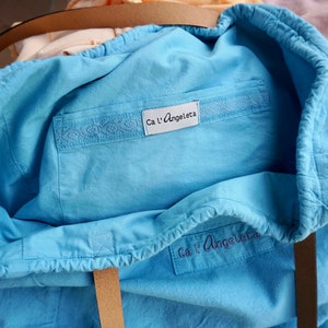 May include: A light blue tote bag with a drawstring closure. The bag has a label that says "Ca l'Angeleta" and a white embroidered design on the front.