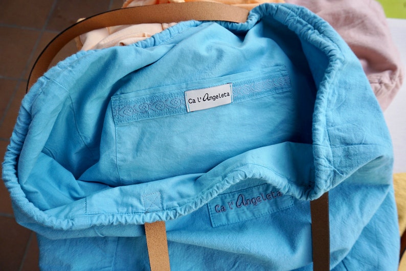 May include: A light blue tote bag with a drawstring closure. The bag has a label that says "Ca l'Angeleta" and a white embroidered design on the front.