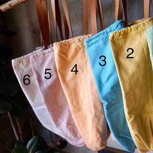 May include: Six canvas tote bags hanging from hooks, each dyed a different pastel color. The bags are numbered 1 through 6, with the numbers visible on the front of each bag.