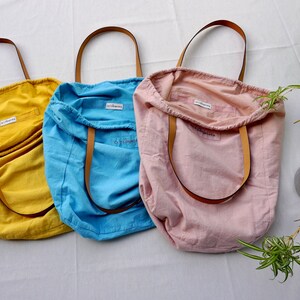 May include: Three tote bags made of natural fabric in yellow, blue, and pink. Each bag has a brown leather handle and a small white tag with the word "Elbowroom" printed on it.