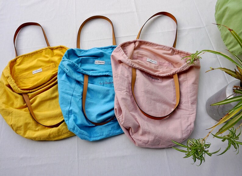 May include: Three tote bags made of natural fabric in yellow, blue, and pink. Each bag has a brown leather handle and a small white tag with the word "Elbowroom" printed on it.