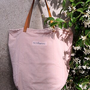 May include: A pale pink canvas tote bag with brown leather straps and the text "Ca l'Angeletta" printed on the front.
