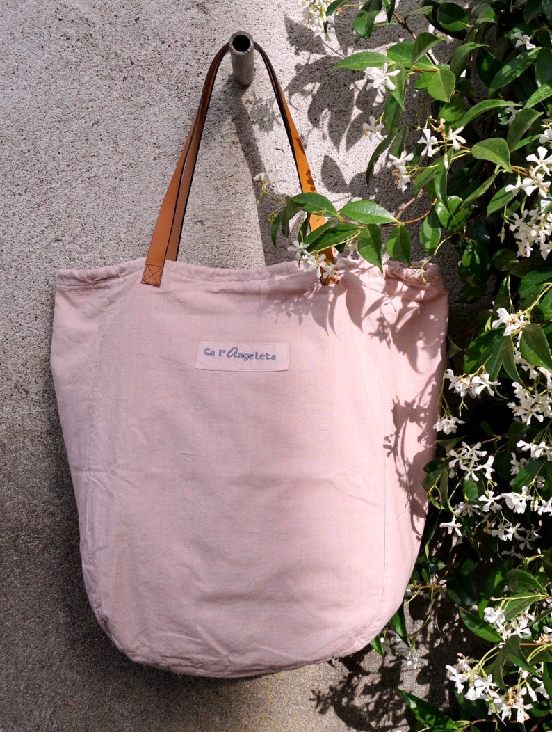 May include: A pale pink canvas tote bag with brown leather straps and the text "Ca l'Angeletta" printed on the front.