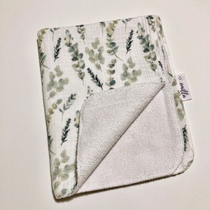 May include: A white muslin baby blanket with a green eucalyptus leaf print on one side and a white terry cloth on the other side. The blanket has a small white tag with the word "omilla" on it.
