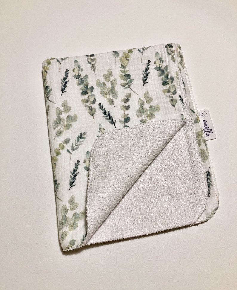 May include: A white muslin baby blanket with a green eucalyptus leaf print on one side and a white terry cloth on the other side. The blanket has a small white tag with the word "omilla" on it.