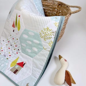 Op de afbeelding: Een patchwork quilt met een thema van de natuur met bomen, huizen en wolken in zachte pastelkleuren. De quilt is opgevouwen in een rieten mand, met een knuffelgans in de buurt. De quilt heeft een lichtblauwe rand.