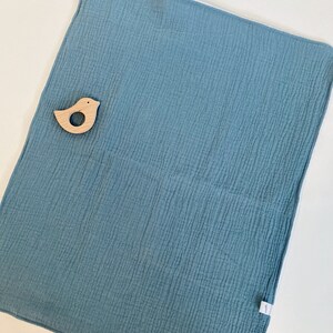 May include: A soft, blue muslin baby blanket with a white border. The blanket is textured and has a small wooden bird toy on top.