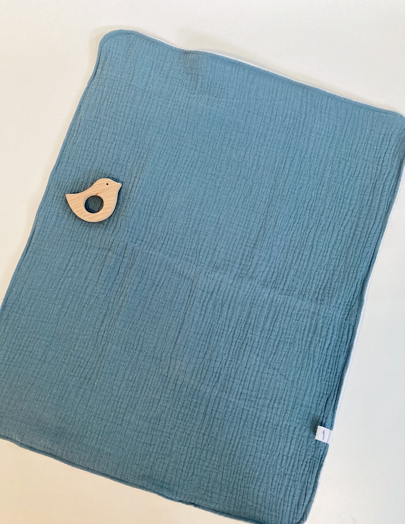 May include: A soft, blue muslin baby blanket with a white border. The blanket is textured and has a small wooden bird toy on top.