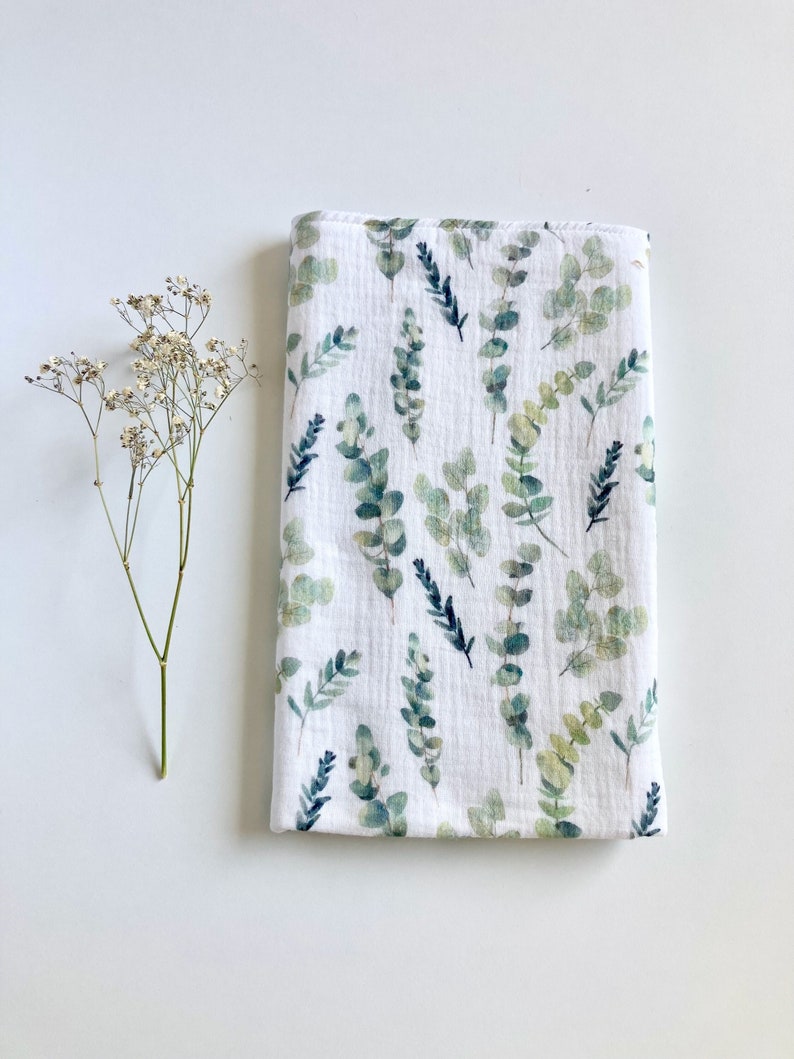 May include: A white muslin swaddle blanket with a green and blue eucalyptus leaf pattern.