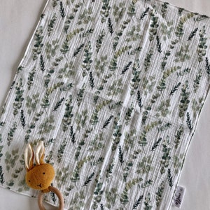 May include: A white muslin baby burp cloth with a green eucalyptus leaf pattern. The cloth has a small tag with the word "Willow" on it. A brown bunny rattle with a wooden ring is on top of the cloth.