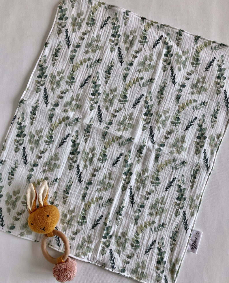 May include: A white muslin baby burp cloth with a green eucalyptus leaf pattern. The cloth has a small tag with the word "Willow" on it. A brown bunny rattle with a wooden ring is on top of the cloth.