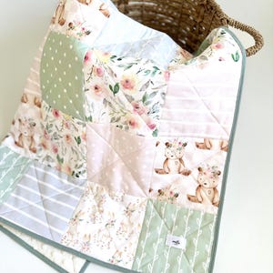 Può includere: Una trapunta patchwork per bambini con una varietà di motivi, tra cui stampe floreali, a pois, a righe e di animali del bosco. La trapunta presenta una morbida tavolozza di colori rosa, verde e bianco, con un bordo verde. La trapunta è piegata in un cestino di vimini.