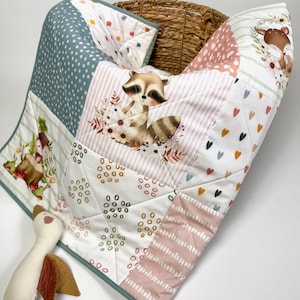Könnte beinhalten: Eine Patchwork-Babydecke mit verschiedenen Mustern und Illustrationen. Die Decke zeigt Waschbär-, Hirsch- und Blumendesigns sowie Punkte, Herzen und Streifen. Die Ränder sind mit einer türkisfarbenen Bordüre versehen, und ein Stoffgans-Spielzeug liegt daneben.