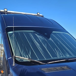 May include: A blue van with a sunshade covering the windscreen. The sunshade is made of a dark material with vertical stripes. The van has a roof rack with lights on top.