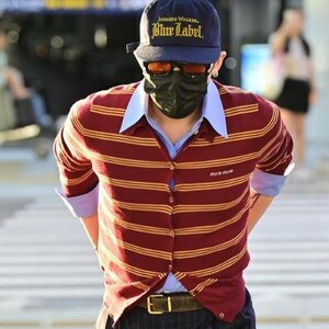 May include: A person wearing a navy blue cap with "Johnnie Walker Blue Label" text, orange sunglasses, and a black face mask. They are wearing a burgundy and gold striped cardigan over a light blue collared shirt, and dark pinstripe pants.