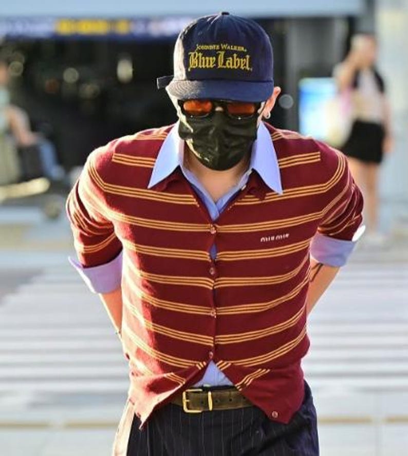 May include: A person wearing a navy blue cap with "Johnnie Walker Blue Label" text, orange sunglasses, and a black face mask. They are wearing a burgundy and gold striped cardigan over a light blue collared shirt, and dark pinstripe pants.
