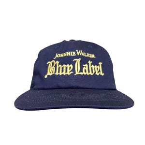 Puede incluir: Gorra de béisbol azul marino con visera curva. La gorra presenta las palabras "JOHNNIE WALKER" sobre "Blue Label" en letras doradas. Un estilo clásico, adecuado para ropa informal.