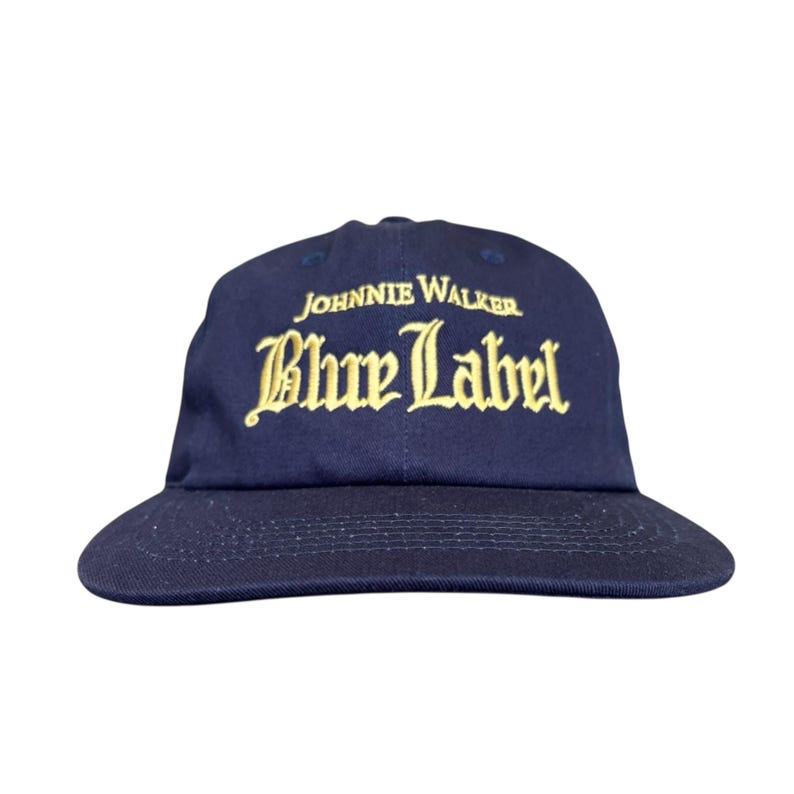 May include: A navy blue baseball cap with a curved brim. The cap features the words "JOHNNIE WALKER" above "Blue Label" in gold lettering. The hat is a classic style, suitable for casual wear.