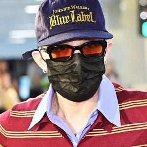 May include: A navy blue baseball cap with "Johnnie Walker Blue Label" text. The person is wearing black sunglasses with orange lenses, a black face mask, and a red and gold striped shirt with a light blue collar.