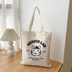 Peut inclure: Un sac fourre-tout en toile blanche avec du texte noir et un chat de dessin animé portant un chapeau. Le texte indique "Genius Lab" avec les années "DIYG 1998" et la phrase "A to the 6, to the U, to STD AKA AGUST D".