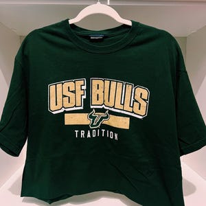 May include: A dark green crop top t-shirt with the text "USF BULLS TRADITION" in gold and white lettering. The shirt is hanging on a white hanger. The shirt is made of a soft material.
