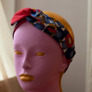 May include: A red, blue and yellow patterned headband with a knot on top. The headband is on a pink mannequin head.