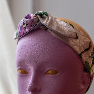 May include: A pink mannequin head wearing a floral headband. The headband is white with a floral pattern in shades of purple, green, and brown.