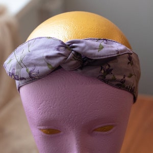 May include: A purple headband with a floral pattern. The headband is tied in a knot on top of the head.
