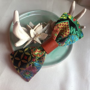 May include: A colourful bow tie with a brown leather strap. The bow tie has a black background with a multi-coloured floral pattern.
