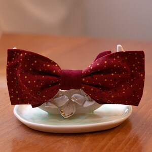 May include: A burgundy bow tie with gold polka dots, displayed on a white ceramic stand.