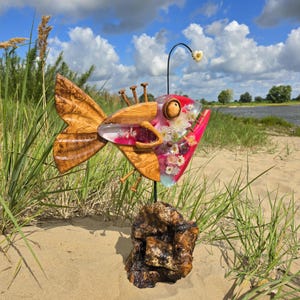 Könnte beinhalten: Eine skurrile Fischskulptur aus Holz und Harz. Der Körper ist mit Blumen gefüllt, der Mund ist rot. Sie steht auf einem Holzsockel und hat eine Blume auf einem Metallstab über sich.