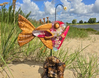 May include: A whimsical wooden fish sculpture with a clear resin body filled with flowers and a red mouth. The fish has wooden fins and is mounted on a wooden base. A black metal rod supports a small flower above the fish.