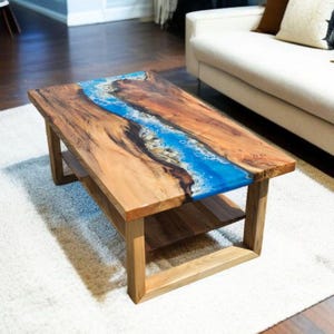 Könnte beinhalten: Rechteckiger Couchtisch aus Holz mit Flussdesign. Der Tisch hat einen hellbraunen Holzrahmen und eine Tischplatte mit einem blauen Epoxidharzfluss. Der Tisch hat ein unteres Regal zur Aufbewahrung. Misst ca. 122 cm lang.