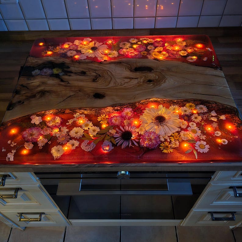May include: A rectangular stove cover board featuring a central piece of natural light-colored wood with prominent grain patterns, flanked by two sections of red resin embedded with various pressed flowers and integrated warm LED fairy lights.