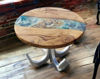 May include: A round wooden table with a blue and white epoxy resin top featuring a river design with seashells and anchors. The table has a silver metal base with three legs shaped like anchors.