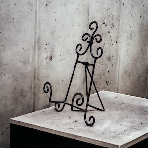 Könnte beinhalten: Ein schwarzer Metall-Staffelei-Ständer mit einem dekorativen Schnörkel-Design. Der Ständer ist aus dünnem Metall gefertigt und hat ein schlichtes, elegantes Design.