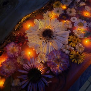 May include: A rectangular wooden board with a live edge, featuring a translucent resin inlay embedded with various pressed flowers including white daisies, purple blossoms, and yellow petals, illuminated by small warm glowing LED lights.