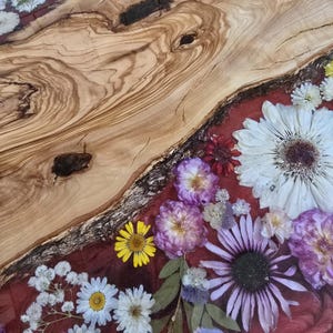 May include: A live edge olive wood board featuring a natural grain pattern with dark knots, paired with a section of red resin embedded with various pressed flowers including large white daisies, purple roses, and small yellow wildflowers.