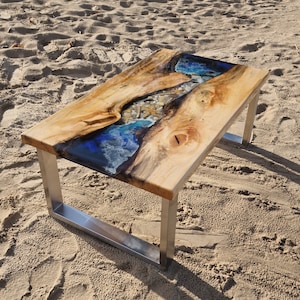 Könnte beinhalten: Ein Holz-Couchtisch mit einer blauen und weißen Epoxidharz-Einlage mit Muscheln. Der Tisch hat einen silbernen Metallrahmen.