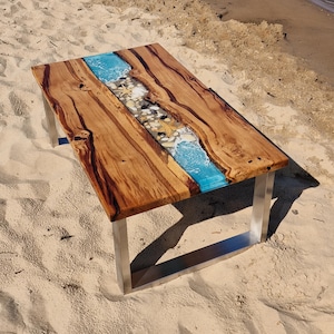 May include: A wooden coffee table with a blue resin river running down the center. The river is filled with white seashells and small white rocks. The table has a stainless steel base.