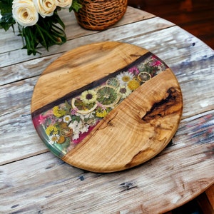 Puede incluir: Una tabla redonda de servir de madera con una incrustación de resina transparente que presenta flores prensadas y rodajas de cítricos secos. La tabla tiene un acabado de madera natural y un aspecto rústico.