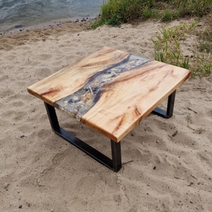 K&ouml;nnte beinhalten: Ein quadratischer Couchtisch mit einer nat&uuml;rlichen Holzplatte und einem schwarzen Metallgestell. Die Platte verf&uuml;gt &uuml;ber einen Fluss aus Epoxidharz mit eingebetteten Muscheln und Seesternen.