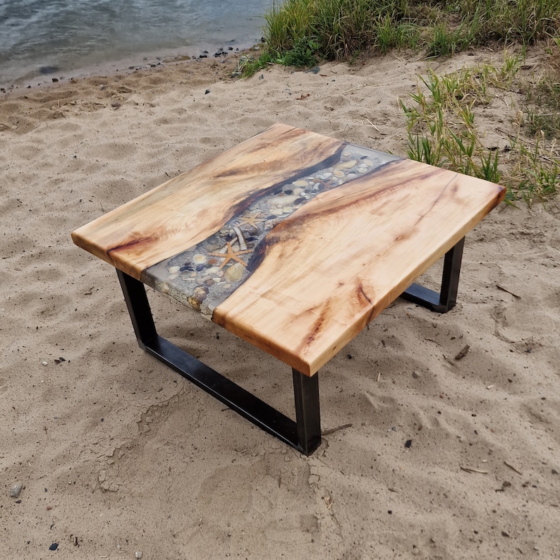 K&ouml;nnte beinhalten: Ein quadratischer Couchtisch mit einer nat&uuml;rlichen Holzplatte und einem schwarzen Metallgestell. Die Platte verf&uuml;gt &uuml;ber einen Fluss aus Epoxidharz mit eingebetteten Muscheln und Seesternen.