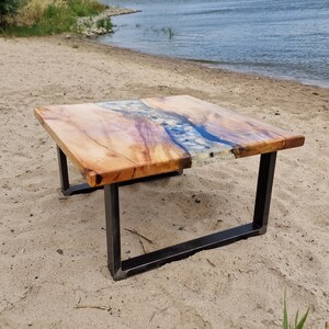 K&ouml;nnte beinhalten: Ein quadratischer Couchtisch mit einer Holzplatte und einem Fluss aus Epoxidharz mit eingebetteten Steinen. Der Tisch hat ein schwarzes Metallgestell.