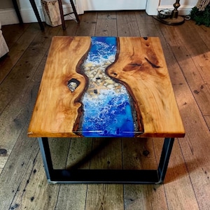 Op de afbeelding: Een rechthoekige salontafel met een houten blad en een blauw hars rivierontwerp. De tafel heeft een zwart metalen onderstel. Het rivierontwerp heeft witte en blauwe wervelingen, die op oceaangolven lijken, met kleine stenen en schelpen.