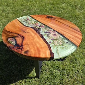 May include: Round wooden table with a green epoxy resin top featuring pressed flowers. The table has a natural wood finish and black metal legs.
