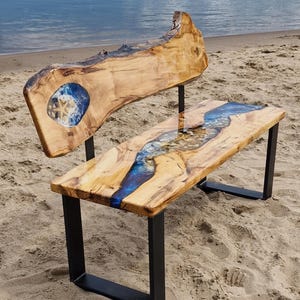 Peut inclure: Banc en bois avec dossier et assise à bord vif, avec un motif de rivière en résine époxy bleue. Le banc a des pieds en métal noir et est posé sur une plage de sable. Le dossier a une incrustation bleue et blanche avec une étoile de mer.