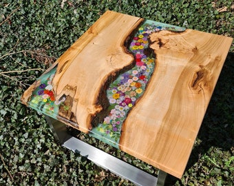 May include: A wooden coffee table with a light wood finish and a resin river filled with pressed flowers. The table has a metal base.