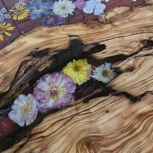 May include: A live edge wood slab table surface featuring a central river of clear resin embedded with various pressed flowers including yellow, white, and purple blossoms. The wood displays prominent natural grain patterns and dark knots.