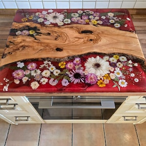 May include: A rectangular kitchen countertop featuring a central live-edge wood slab flanked by two sections of deep red resin embedded with a variety of pressed dried flowers, including white daisies, purple asters, and green fern leaves.